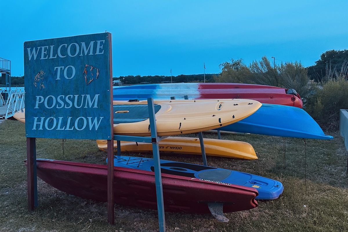 Kayaks and paddleboards lined up by the Welcome to Possum Hollow sign at dusk